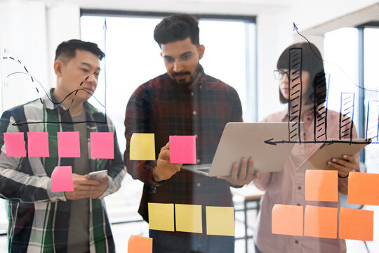 Team members collaborating in brainstorming session with sticky notes on glass wall analyzing charts and data diagrams. Clear focus on teamwork and productivity in modern office environment.