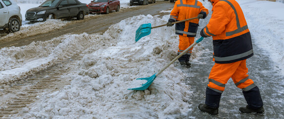 Two men in orange and black work together to clear snow from a street