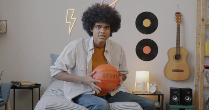 Portrait of African American teenage boy watching basketball competition on TV sitting in bedroom alone looking at camera. Sports and fans concept.