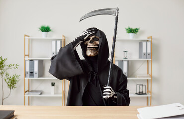 Stressed man in grim reaper death costume with black cloak and scythe sitting at desk and putting...