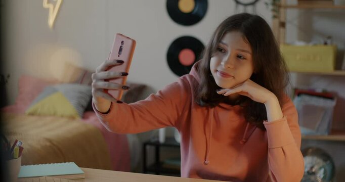 Cute teenage girl posing for smartphone camera taking selfie indoors in bedroom in apartment. Photograph and youth concept.