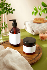 The image showcases an amber glass pump bottle and a black jar with blank labels, surrounded by a wooden cutting board, a white towel, pink salt bowl, and greenery, perfect for beauty product ads.