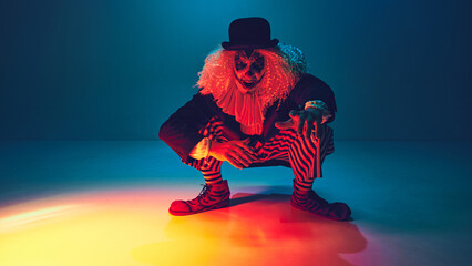 Crouched with menace, clown dressed retro clothes reaches forward, in neon light against gradient background. Concept of Happy Halloween, party, festive mood, celebration. Ad