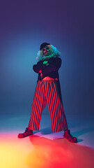 Tall and confident, clown crosses his arms and looking at camera in neon light against gradient background. Concept of Happy Halloween, party, festive mood, celebration. Ad