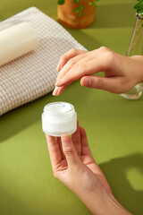 Spa and beauty product advertisement featuring a female hand gently opening a white cream jar, revealing its smooth texture, surrounded by a towel and a potted plant. Sunlight casts a warm the scene.