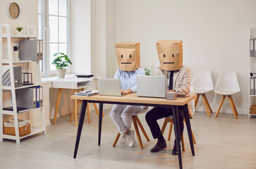 Two funny faceless company employees wearing paper bags on head with drawn upset faces sitting at desk on workplace with laptops. Anonymous business people man and woman working in office
