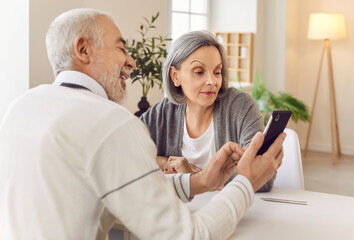 Portrait of an elderly senior couple sitting at the table in living room at home and using smartphone together browsing apps. Senior man showing video or news on mobile phone to his wife.