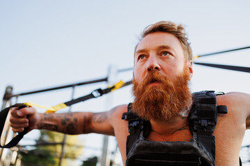 Obraz premium Portrait of bearded sportsman training with suspension straps in park