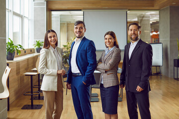 Portrait of a happy young business people men and women looking at camera and smiling indoors. Company employees or group of staff standing in office in suits confidently and cheerfully.