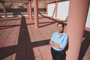 Confident woman in blue shirt standing outdoors on a sunny day, leaning against a pink column with shadows on the ground