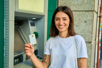 Smiling young woman using an ATM on a city street, showing her card, with a green handbag.