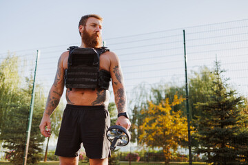 Muscular sportsman in weighted vest holding ab roller outdoors