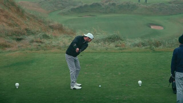 Male golfer with backwards white hat hits ball off tee from white tees on par three on a rainy foggy day on Ireland links golf course as other golfers watch on in slow motion