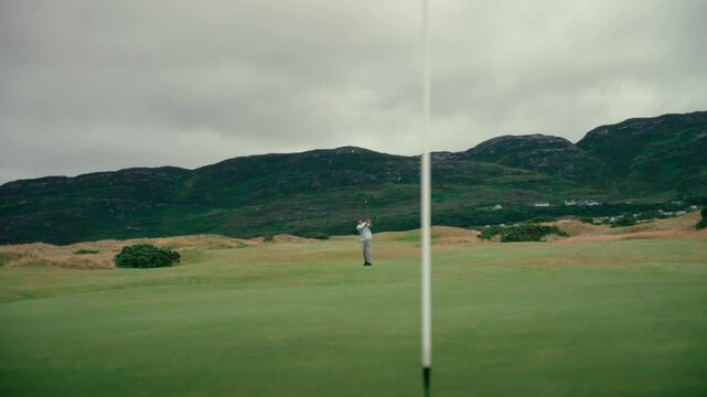 Male golfer hits chip shot onto putting green on Ireland Links golf course, ball bounces in slow motion and rolls away from pin