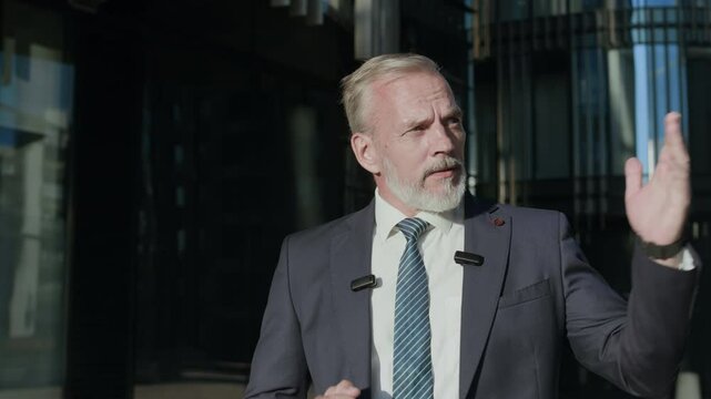 Tracking medium shot of confident Caucasian adult man in blue suit with two small wireless microphones attached to lapel covering business news during video report recording in city downtown