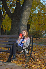 a girl sits on a bench in an autumn park, headphones on her head, a smartphone in her hands and a cup with a warm drink: tea, coffee or cappuccino. hobbies, recreation and entertainment