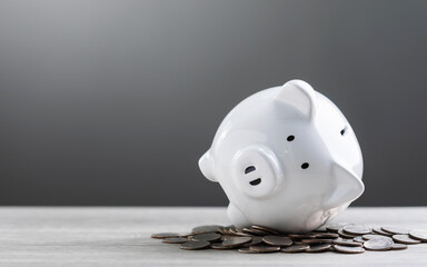 White piggy bank and a pile of coins on a wooden table against a grey background, with ample copy space for text. The image symbolizes savings, financial planning, and responsible money management.