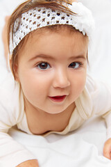 Tiny tot in cute outfit peeks over blanket on soft bed with white bedding, smiling happily, her face full of joy against studio background. Concept of beauty, parenthood, childhood, life and birth.