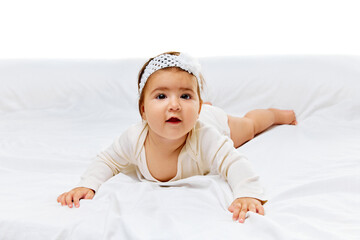 Sweet girl in white onesie and floral headband gazes up, resting comfortably on soft bed against white studio background. Concept of beauty, parenthood, childhood, life and birth.