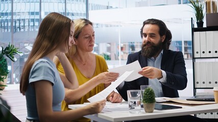 Financial advisor talks with clients about importance of budgeting in income management, offering them written guide. Mom and daughter explore best practices for building money saving habits, camera A