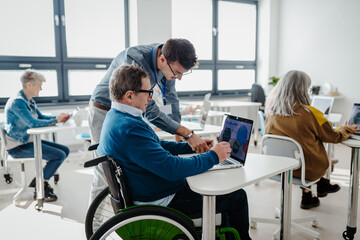 Teacher explaining senior man on wheelchair how to work with laptop. Elderly people attending computer and technology education class. Digital literacy.