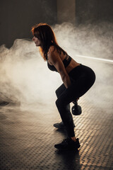 A focused young woman is performing a kettlebell exercise in a gym that features dramatic lighting,...