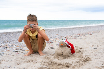 Child playing on the seashore with a snowman in a Santa hat made of beach sand. Christmas in summer