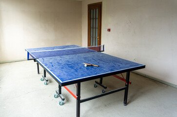 Blue table tennis table covered with dust with a stretched net with a red racket and ball in an empty bright room