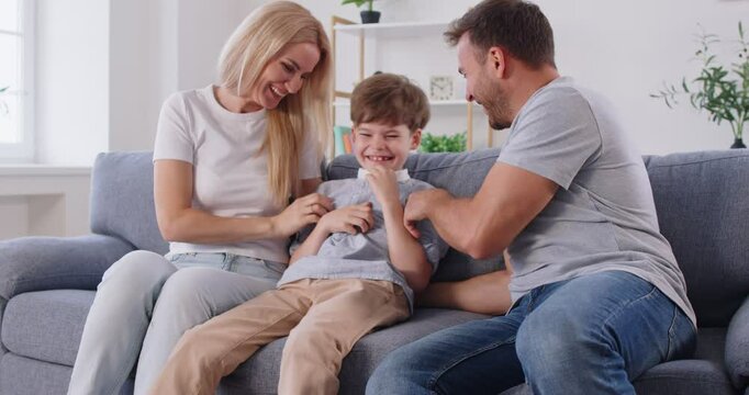 Happy family, cheerful people sitting on couch in living room to have fun, little son tickling mother, laughing together with parents, enjoy free time playing, overjoyed fun at home on weekend