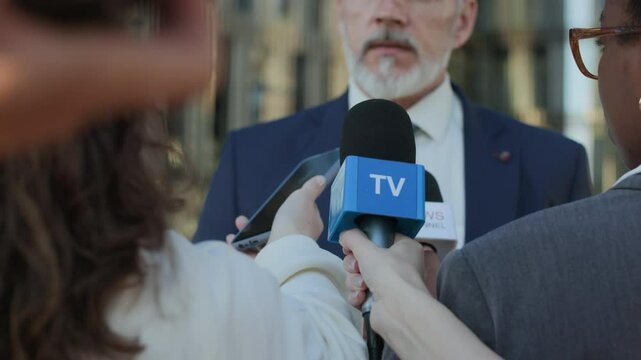 Middle-aged Caucasian man in politics wearing formal suit and tie answering reporters questions and making speech while being interviewed outdoors after press conference