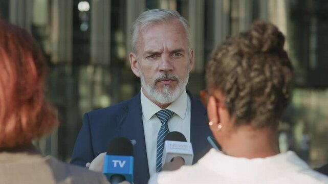 Waist up shot of confident mature Caucasian male politician wearing blue suit, white shirt and blue striped tie giving interview to TV reporters while standing outdoors