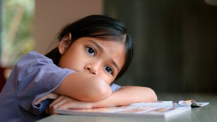 Naklejka premium An Asian girl sits at a table in a library, her face clearly showing boredom as she looks away from the open book in front of her. She looks disinterested and lost in thought.