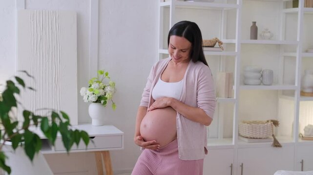 Happy Caucasian middle aged woman warmly caressing a pregnant belly at home living room. Concept of a pregnancy, expectation and child affectionate.