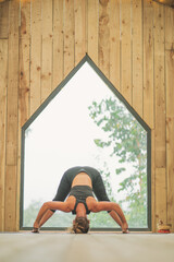 Woman doing Vinyasa yoga pose close to a window with black fitness outfit wooden room natural light wide leg forward pose D