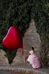 A young lady in a pink princess dress poses on the street and holds a huge red ball in the shape of a heart, a girll walks along the street of an ancient city. Concept of a postcard for February 14