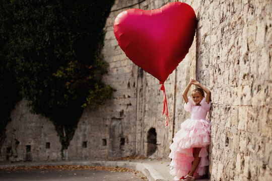A young lady in a pink princess dress poses on the street and holds a huge red ball in the shape of a heart, a girll walks along the street of an ancient city. Concept of a postcard for February 14