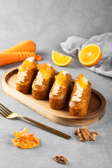 Appetizing carrot-orange cupcakes with cream and carrot chips lie on a wooden plate with carrots, orange and nuts on a gray background. Baking concept for a coffee shop.
