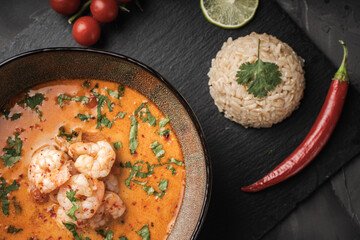 Tom Yam Kung Spicy Thai soup with shrimp in a bowl on a dark background and red pepper, cilantro rice on a black mica board. top view