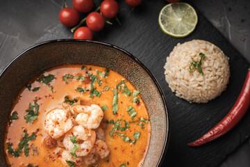 Tom Yum Kung Spicy Thai soup with shrimp in a bowl on a dark background and red pepper, rice and lime with cilantro and chopsticks on a black mica board. Copyspace