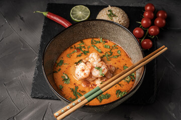Tom Yum Kung Spicy Thai soup with shrimp in a bowl on a dark background and red pepper, rice and lime with cilantro and chopsticks on a black mica board. Copyspace