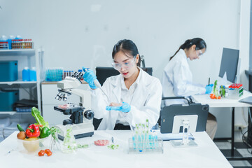 Two Asian women in a lab work on food research, using a microscope, petri dish, and test tubes...