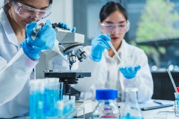 Two Asian women in a lab work on food research, using a microscope, petri dish, and test tubes filled with chemical solutions. They study vegetables, pork, and plants for GMO traits and nutrition.