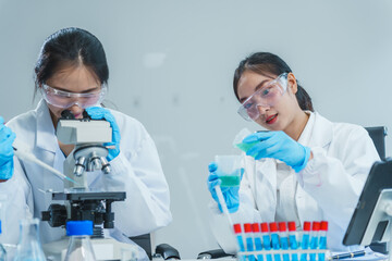 Two Asian women in a lab work on food research, using a microscope, petri dish, and test tubes filled with chemical solutions. They study vegetables, pork, and plants for GMO traits and nutrition.