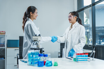 Shaking hands Two Asian women in lab work on food research, using microscope, petri dish, test tubes filled with chemical solutions. They study vegetables, pork, plants for GMO traits and nutrition.