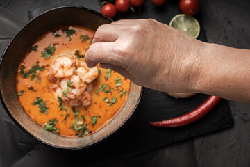 Hand sprinkles Tom Yum Kung seasoning Spicy Thai soup with shrimp in a bowl on a dark background and red pepper, rice and lime with cilantro and chopsticks on a black mica board.