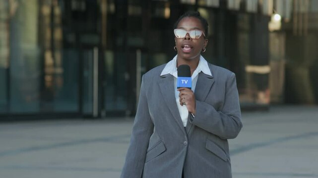 Medium shot of confident young Black female TV reporter in grey suit walking towards camera with microphone while covering news in downtown, with glass building on blurred background