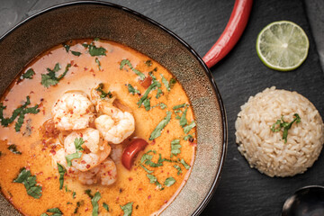 Tom Yam Kung Spicy Thai soup with shrimp in a bowl on a dark background and red pepper, rice and lime with cilantro and a spoon on a black mica board.
