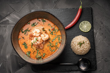 Tom Yam Kung Spicy Thai soup with shrimp in a bowl on a dark background and red pepper, rice and lime with cilantro and a spoon on a black mica board.