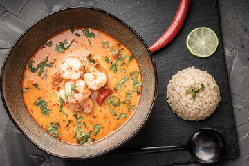 Tom Yam Kung Spicy Thai soup with shrimp in a bowl on a dark background and red pepper, rice and lime with cilantro and a spoon on a black mica board.