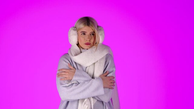 Portrait Of Young Blonde Woman Feeling Cold, Wearing Scarf Grey Sweater and Earmuffs, Studio Shot With Pink Background